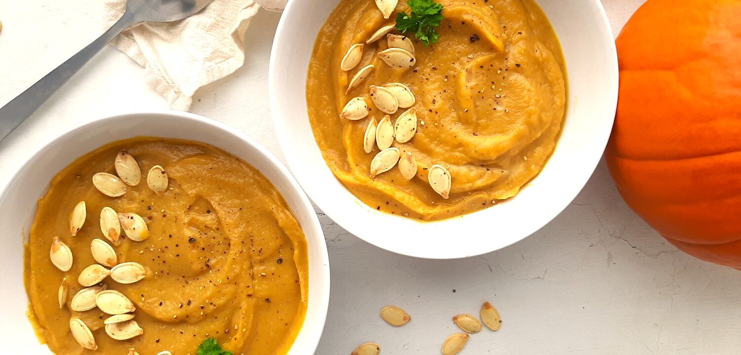 Two bowls of hearty roast pumpkin soup from scratch in white bowls, garnished with pumpkin seeds, fresh parsley sprigs, and fresh cracked pepper. A whole sugar pie pumpkin, beige cloth, silver spoon, and more pumpkin seeds are scattered around the white background. Sugar with Spice Blog.