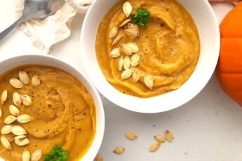 Two bowls of hearty roast pumpkin soup from scratch in white bowls, garnished with pumpkin seeds, fresh parsley sprigs, and fresh cracked pepper. A whole sugar pie pumpkin, beige cloth, silver spoon, and more pumpkin seeds are scattered around the white background. Sugar with Spice Blog.