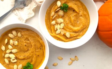 Two bowls of hearty roast pumpkin soup from scratch in white bowls, garnished with pumpkin seeds, fresh parsley sprigs, and fresh cracked pepper. A whole sugar pie pumpkin, beige cloth, silver spoon, and more pumpkin seeds are scattered around the white background. Sugar with Spice Blog.