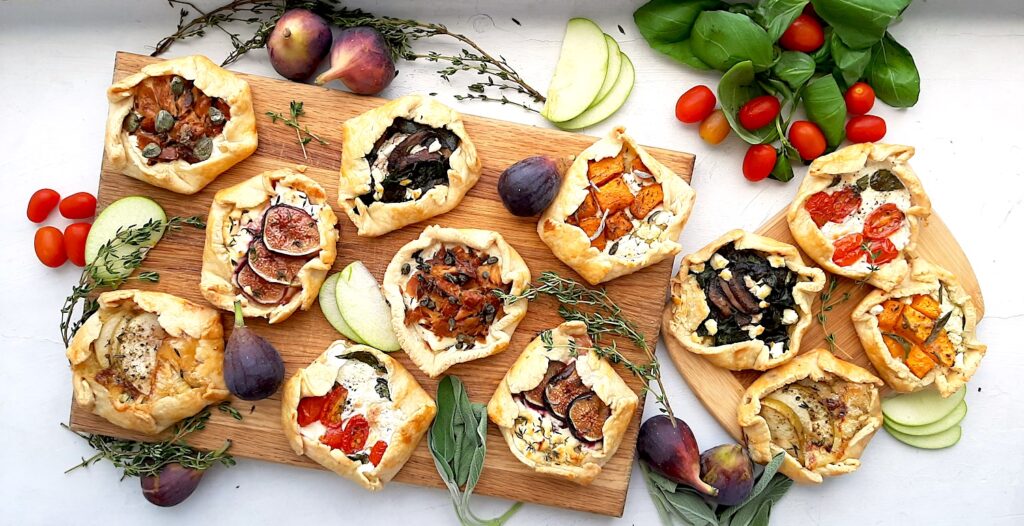 Assorted mini savory galettes on one large and one small wooden cutting board surrounded by fresh herbs, vegetables, and fruits on a white background. Overhead shot. Sugar with Spice Blog.
