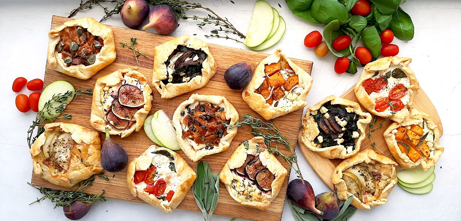 Assorted mini savory galettes on one large and one small wooden cutting board surrounded by fresh herbs, vegetables, and fruits on a white background. Overhead shot. Sugar with Spice Blog.