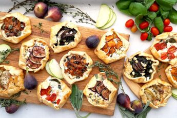 Assorted mini savory galettes on one large and one small wooden cutting board surrounded by fresh herbs, vegetables, and fruits on a white background. Overhead shot. Sugar with Spice Blog.