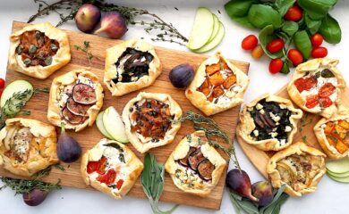 Assorted mini savory galettes on one large and one small wooden cutting board surrounded by fresh herbs, vegetables, and fruits on a white background. Overhead shot. Sugar with Spice Blog.