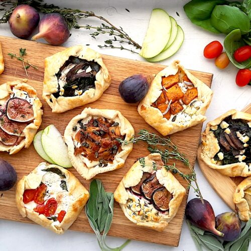 Assorted mini savory galettes on one large and one small wooden cutting board surrounded by fresh herbs, vegetables, and fruits on a white background. Overhead shot. Sugar with Spice Blog.