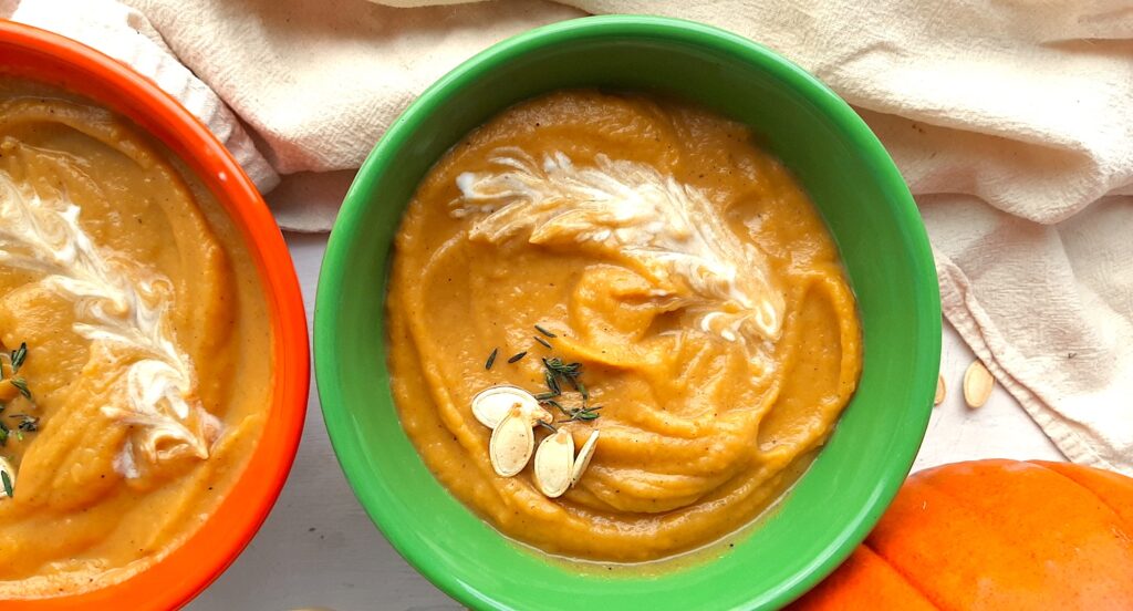 A bowls of hearty roast pumpkin soup from scratch in a green bowl, garnished with pumpkin seeds, fresh parsley sprigs, and coconut cream. A second bowl of soup in an orange bowl, whole sugar pie pumpkin, beige cloth, silver spoon, and more pumpkin seeds are scattered around the white background. Sugar with Spice Blog.