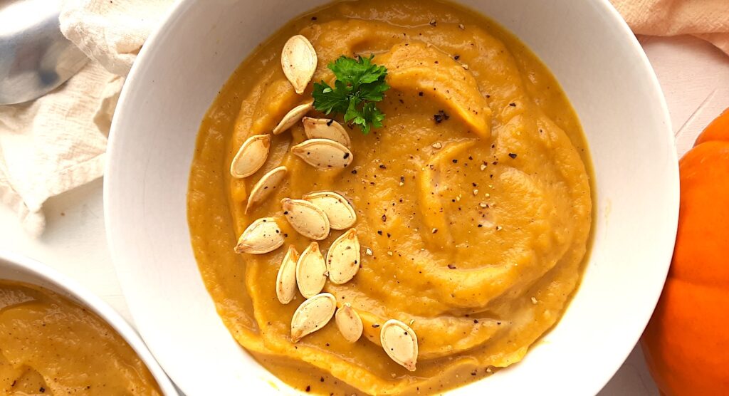 A bowl of hearty roast pumpkin soup from scratch in a white bowl, garnished with pumpkin seeds, fresh parsley sprigs, and fresh cracked pepper. A whole sugar pie pumpkin, beige cloth, silver spoon, a second bowl of soup, and more pumpkin seeds are scattered around the white background. Close up shot. Sugar with Spice Blog.