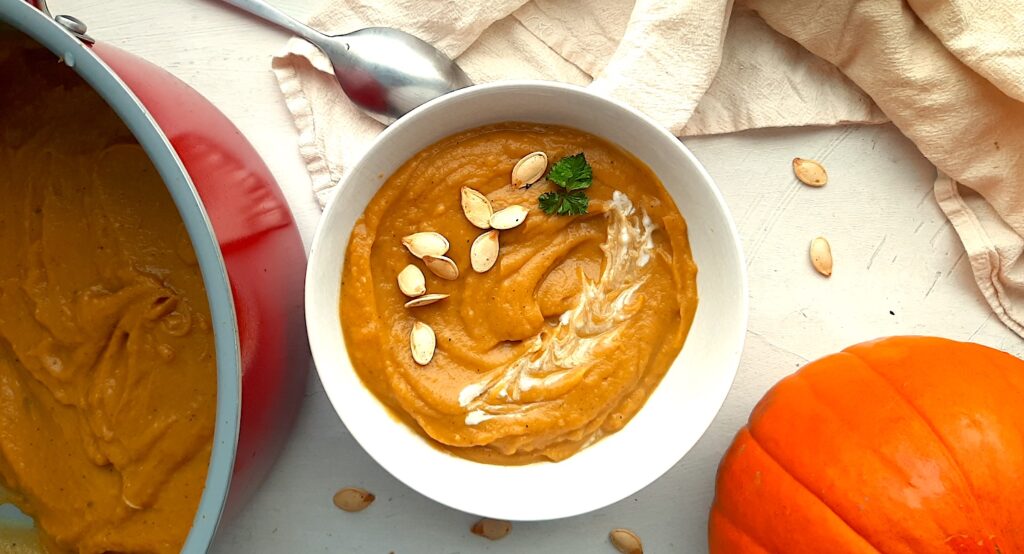 A bowl of hearty roast pumpkin soup from scratch in a white bowl, garnished with pumpkin seeds, fresh parsley sprigs, and coconut cream. A red soup pot with more pumpkin soup to the side. A whole sugar pie pumpkin, beige cloth, silver spoon, and more pumpkin seeds are scattered around the white background. Sugar with Spice Blog.