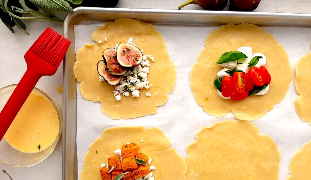 Raw mini savory galettes being assembled on a parchment paper-lined baking tray. Six small pie crust rounds with three filled with fig, tomato, and butternut squash. Three others empty. Egg wash in a pyrex dish with a red pastry brush to the side. Fresh figs and sage to the side. White Background overhead shot. Sugar with Spice Blog