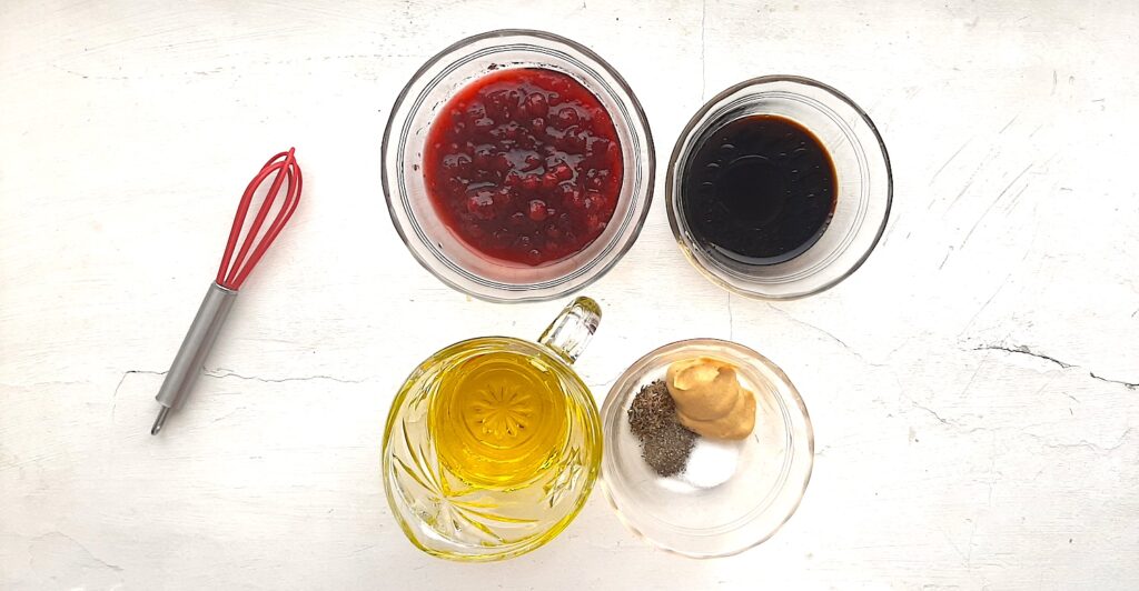 Ingredients for cranberry balsamic dressing. Glass pitcher of olive oil, three pyrex dishes with cranberry sauce, balsamic vinegar, dried thyme, pepper, salt, and dijon mustard. A small red silicone whisk nearby. All on a white background. Sugar with Spice Blog.