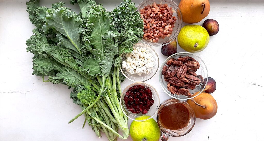 Ingredients for winter pear salad. Fresh bunch of kale, two fresh bosc pears, two fresh d'anjou pears, three fresh mission figs. Sautéed pancetta, fresh gorgonzola, maple candied pecans, dried cranberries all in pyrex bowls. Small glass pitcher of cranberry balsamic dressing. All on a white background. Overhead shot. Sugar with Spice Blog.