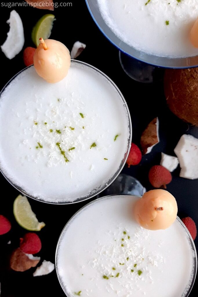 Coconut lychee martinis in three martini glasses, garnished with shaved coconut, lime zest, and a canned lychee. Fresh coconut, lime wedges, and lychees surround on a black background. Overhead shot. Sugar with Spice Blog. Pinterest image.