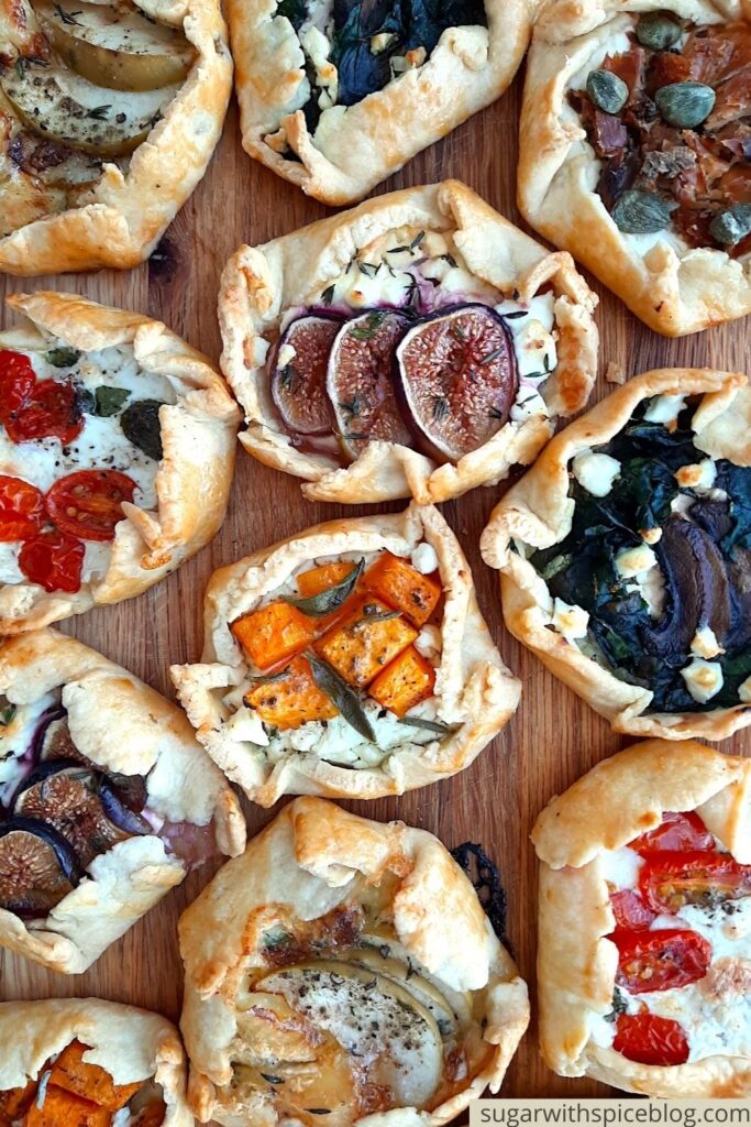 Assorted mini savory galettes on a wooden cutting board. Overhead shot. Sugar with Spice Blog. Pinterest Image.