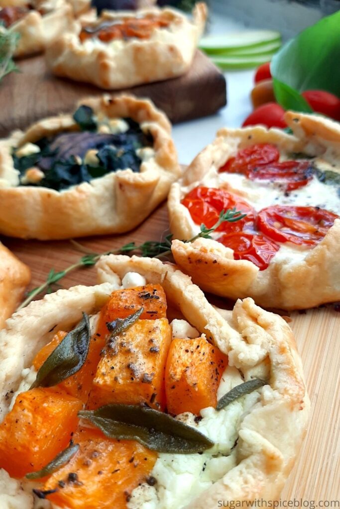 Mini Savory Galette of butternut squash, goat cheese, and sage on a wooden cutting board with assorted mini savory galettes and fresh thyme and tomatos behind. Overhead front shot. Sugar with Spice Blog. Pinterest Image.