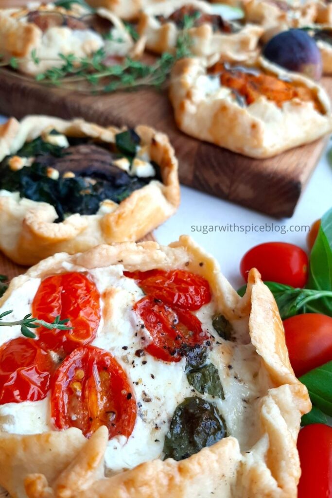 Mini Savory Galette of caprese tomato, basil, and mozzarella on a wooden cutting board with assorted mini savory galettes, fresh basil, tomatoes, and fresh thyme behind. Overhead front shot. Sugar with Spice Blog. Pinterest Image.