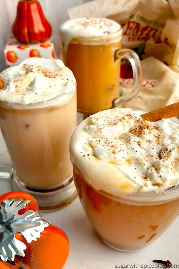 Three glasses of pumpkin spice hot buttered rum topped with whipped cream, cinnamon sticks, and ground nutmeg on a white windowsill. Window sill decorated with ceramic pumpkins, whole cloves, whole cinnamon sticks, ground nutmeg, and orange or fall-themed dish towels. Front Shot. Sugar with Spice blog. Pinterest Image.