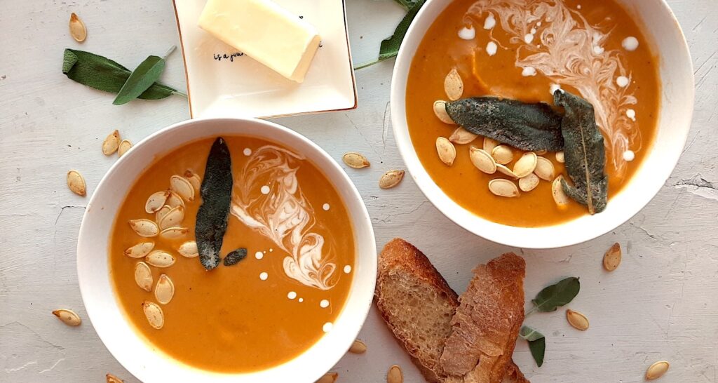 Brown Butter butternut squash soup in two white bowls garnished with roast squash seeds, fried sage, and cream swirls. More sage leaves, roast squash seeds scatter around the white background. Pat of butter on a cream ceramic dish and two slices of sourdough bread nearby. Overhead shot. Sugar with Spice Blog.