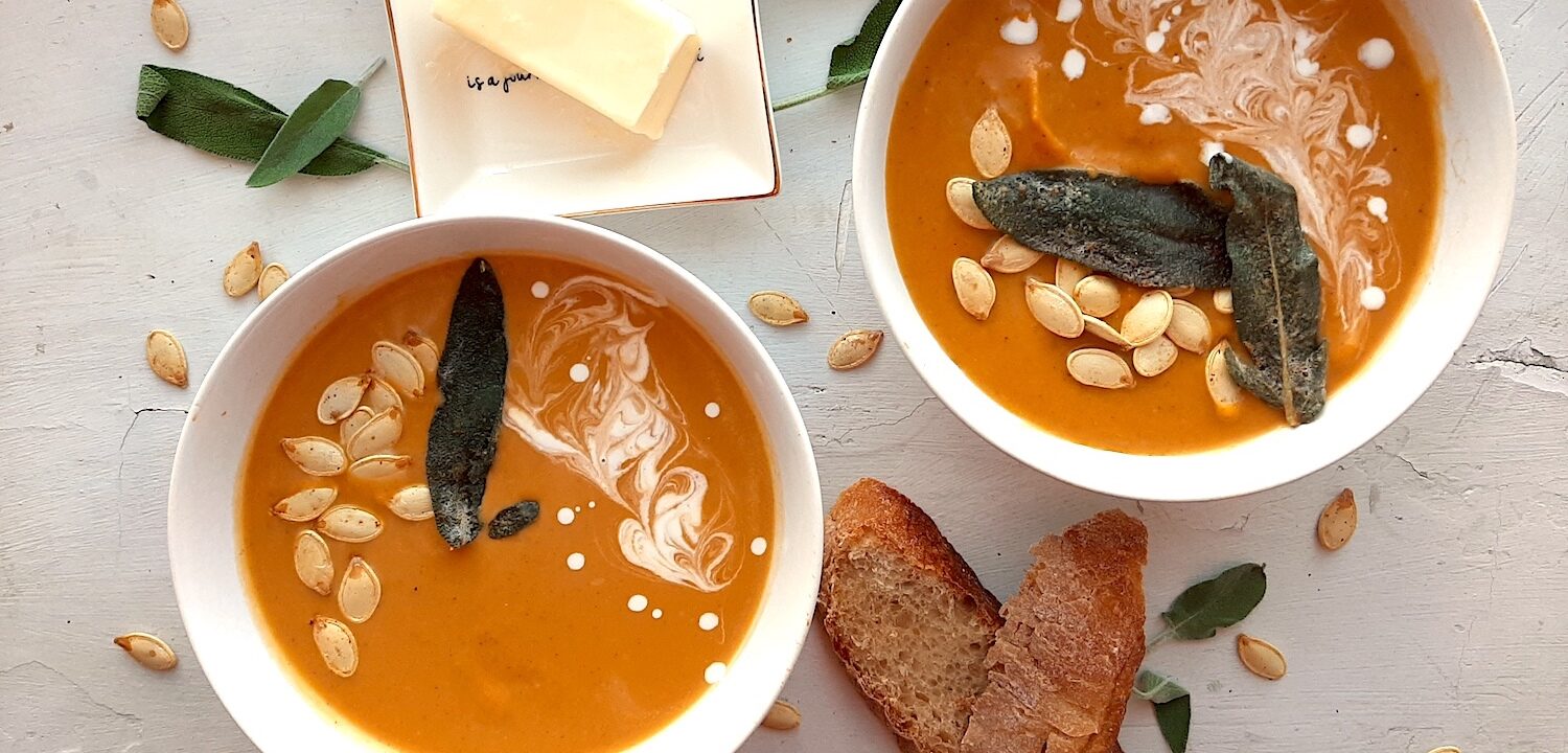 Brown Butter butternut squash soup in two white bowls garnished with roast squash seeds, fried sage, and cream swirls. More sage leaves, roast squash seeds scatter around the white background. Pat of butter on a cream ceramic dish and two slices of sourdough bread nearby. Overhead shot. Sugar with Spice Blog.