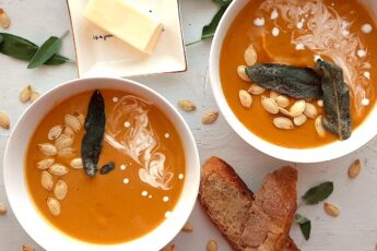 Brown Butter butternut squash soup in two white bowls garnished with roast squash seeds, fried sage, and cream swirls. More sage leaves, roast squash seeds scatter around the white background. Pat of butter on a cream ceramic dish and two slices of sourdough bread nearby. Overhead shot. Sugar with Spice Blog.