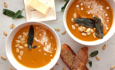 Brown Butter butternut squash soup in two white bowls garnished with roast squash seeds, fried sage, and cream swirls. More sage leaves, roast squash seeds scatter around the white background. Pat of butter on a cream ceramic dish and two slices of sourdough bread nearby. Overhead shot. Sugar with Spice Blog.