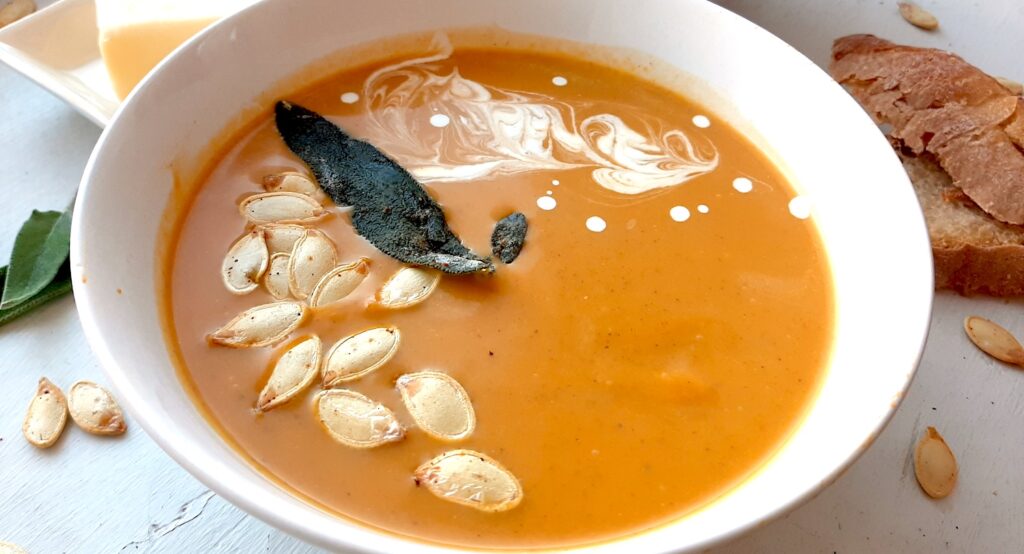 Brown Butter butternut squash soup in a white bowl garnished with roast squash seeds, fried sage, and cream swirls. More sage leaves, roast squash seeds scatter around the white window sill. Pat of butter on a cream ceramic dish and two slices of sourdough bread nearby. Close up side shot. Sugar with Spice Blog.