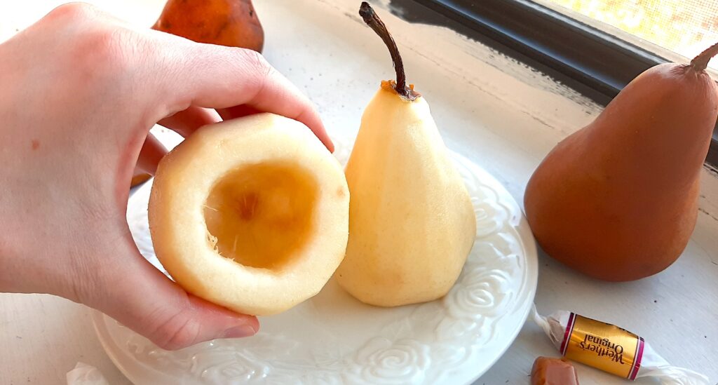 Two bosc pears, peeled and cored. A woman's hand holds up one pear to show the core removed. Two more bosc pears nearby. Caramel candies nearby. All on a white windowsill. Sugar with Spice Blog.