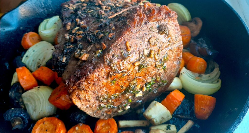 Bottom Round Roast Beef with garlic herb butter rub on a layer of carrots, parsnips, mushrooms, and onions in a large cast iron skillet. Sugar with Spice Blog.
