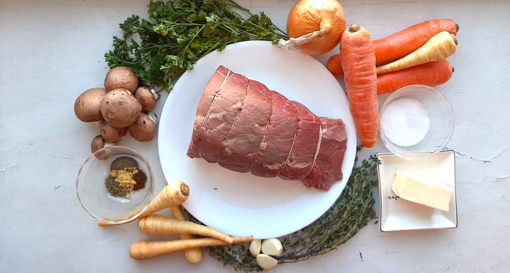 Ingredients for a garlic herb bottom round roast beef. Raw bottom round roast on a white plate surrounded by whole carrots, whole parsnips, whole white onion, whole mushrooms, whole garlic cloves, fresh parsley, fresh thyme, a pat of raw butter, kosher salt in a pyrex bowl, and pyrex bowl with marjoram, pepper, cloves, and ground mustard. All on a white background. Sugar with Spice Blog.