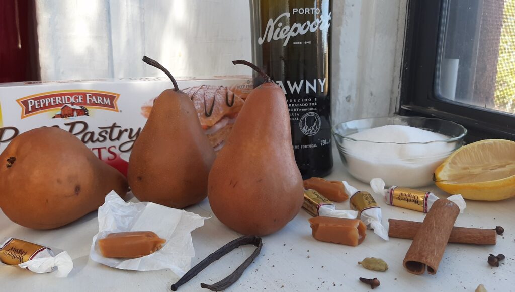 Ingredients for port-poached pears in puff pastry with caramel filling. Bosc pears, puff pastry shell box, tawny port bottle, pyrex bowl of granulated sugar, sliced lemon, whole vanilla bean, cinnamon sticks, cardamom pods, whole cloves, ground nutmeg, and caramel candies all on a white window sill. Sugar with Spice Blog.
