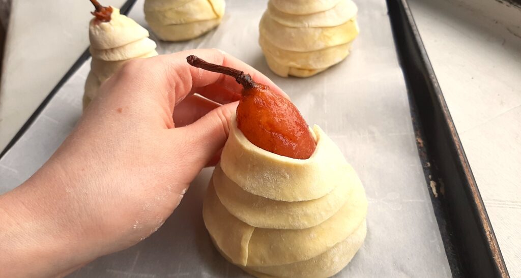 Port-poached pears being wrapped in puff pastry. A port-poached pear on a baking sheet. A woman's hand wraps puff pastry strips around the pear. Three other pears already wrapped sit behind on the rest of the baking sheet. All on a white window sill. Sugar with Spice Blog.