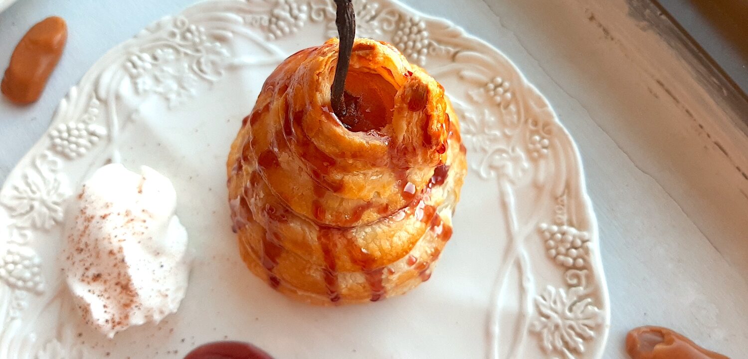 Port poached pear wrapped in puff pastry and drizzled with tawny port sauce on a cream ceramic plate. A side of whipped cream sprinkled with nutmeg, a streak of port sauce on the plate. Caramel candies nearby. On a white window sill. Sugar with Spice Blog.