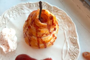 Port poached pear wrapped in puff pastry and drizzled with tawny port sauce on a cream ceramic plate. A side of whipped cream sprinkled with nutmeg, a streak of port sauce on the plate. Caramel candies nearby. On a white window sill. Sugar with Spice Blog.