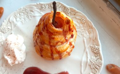 Port poached pear wrapped in puff pastry and drizzled with tawny port sauce on a cream ceramic plate. A side of whipped cream sprinkled with nutmeg, a streak of port sauce on the plate. Caramel candies nearby. On a white window sill. Sugar with Spice Blog.