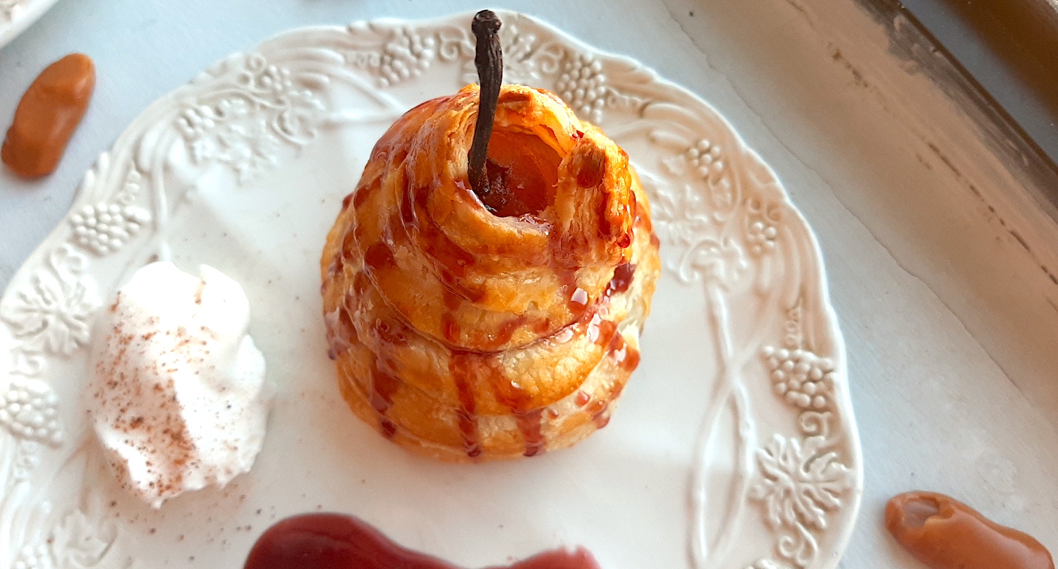 Port poached pear wrapped in puff pastry and drizzled with tawny port sauce on a cream ceramic plate. A side of whipped cream sprinkled with nutmeg, a streak of port sauce on the plate. Caramel candies nearby. On a white window sill. Sugar with Spice Blog.