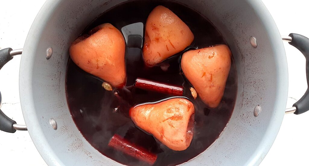 Four peeled and cored bosc pears poaching in tawny port wine with cinnamon sticks, cloves, and cardamom pods. All in a soup pot on a white window silly, overhead shot. Sugar with Spice Blog.