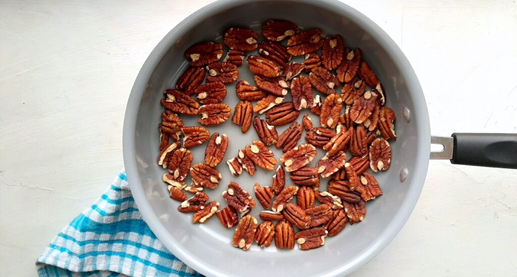 Maple candied pecans being toasted in a grey sauté pan with a blue checked dish cloth near by. White background. Sugar with Spice Blog.