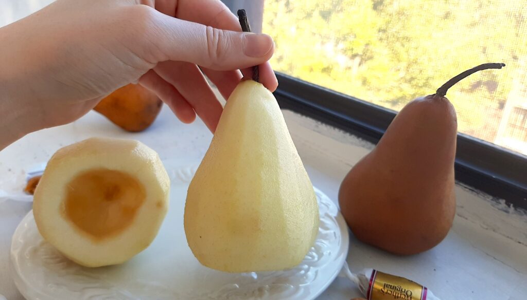 Two bosc pears, peeled and cored. A woman's hand holds up one pear to show the pealed skin. Cored and peeled pear rests behind, cored side up, on a plate. Two more bosc pears nearby. Caramel candies nearby. All on a white windowsill. Sugar with Spice Blog.