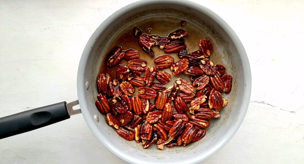 Maple candied pecans phase 2 when maple syrup is still liquid. Pecans, maple syrup, cinnamon, nutmeg, and salt in a grey sautépan on a white background. Sugar with Spice Blog.