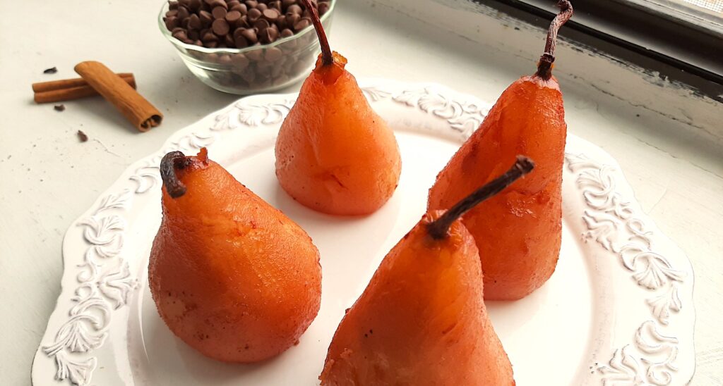 Tawny port-poached pears cooling on a decorated white ceramic plate. Cinnamon sticks, cloves, and a pyrex bowl of milk chocolate chips nearby. All on a white window sill. Sugar with Spice Blog.