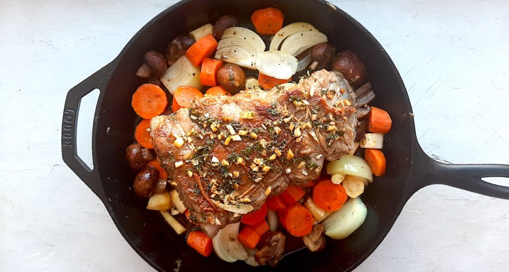 Bottom Round Roast Beef with garlic herb butter rub on a layer of carrots, parsnips, mushrooms, and onions in a large cast iron skillet. White background, overhead shot. Sugar with Spice Blog.