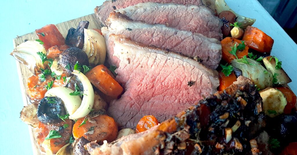 Garlic and Herb Bottom Round Roast Beef on a cutting board, several medium rare slices displayed. Surrounded by roasted carrots, parsnips, onions, and mushrooms. Sprinkled with fresh parsley. On a white background. Sugar with Spice Blog.