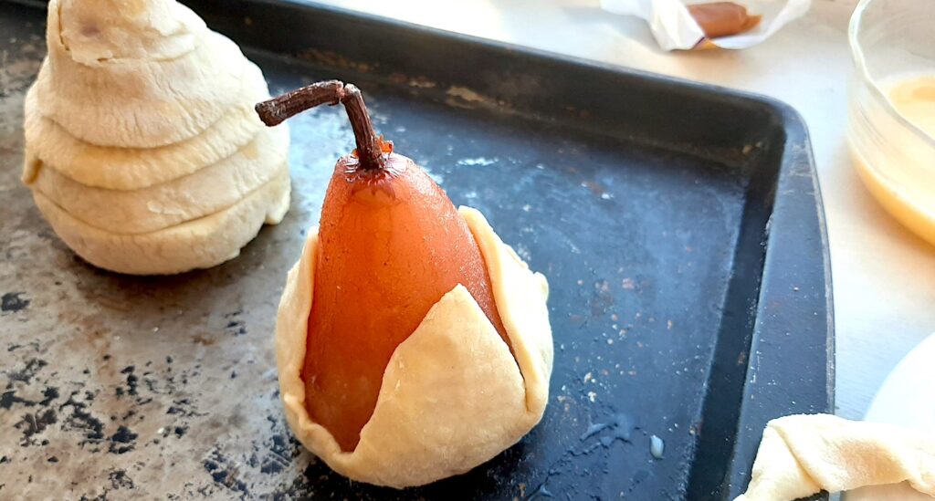 Port poached pear being sealed with puff pastry. A second pear already wrapped in puff pastry sits behind on the baking sheet. Pyrex bowl of egg wash, caramel candies, and extra pastry strips nearby. Sugar with Spice Blog.