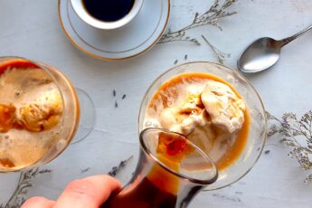 Earl Grey tea affogato preparation. A woman's hand pours a carafe of espresso over a glass coupe with earl grey tea ice cream. A second coupe with earl grey tea ice cream and espresso and a white espresso cup with more espresso to one side. A silver spoon, lavender sprigs, and babies breath flowers scattered around on the white background. Sugar with Spice Blog.