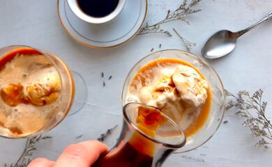 Earl Grey tea affogato preparation. A woman's hand pours a carafe of espresso over a glass coupe with earl grey tea ice cream. A second coupe with earl grey tea ice cream and espresso and a white espresso cup with more espresso to one side. A silver spoon, lavender sprigs, and babies breath flowers scattered around on the white background. Sugar with Spice Blog.