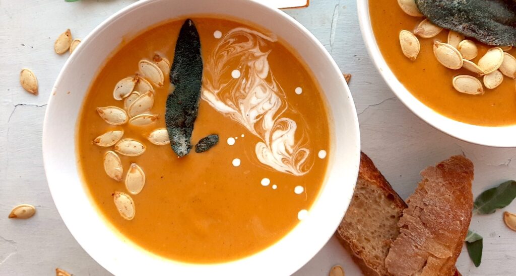 Brown Butter butternut squash soup in a white bowl garnished with roast squash seeds, fried sage, and cream swirls. A second bowl to one side. More sage leaves, roast squash seeds scatter around the white background. Pat of butter on a cream ceramic dish and two slices of sourdough bread nearby. Overhead shot. Sugar with Spice Blog.