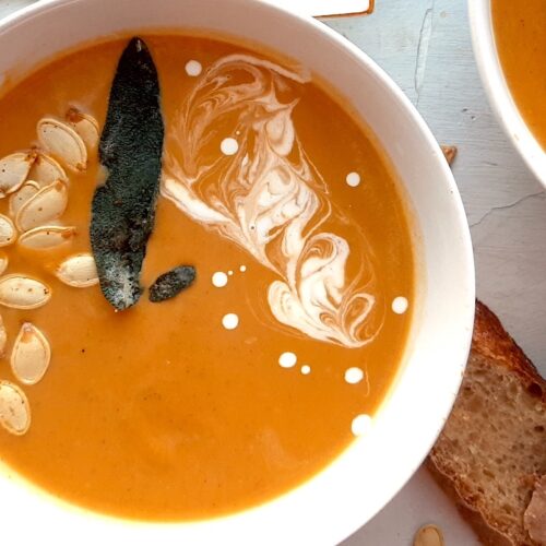 Brown Butter butternut squash soup in a white bowl garnished with roast squash seeds, fried sage, and cream swirls. A second bowl to one side. More sage leaves, roast squash seeds scatter around the white background. Pat of butter on a cream ceramic dish and two slices of sourdough bread nearby. Overhead shot. Sugar with Spice Blog.