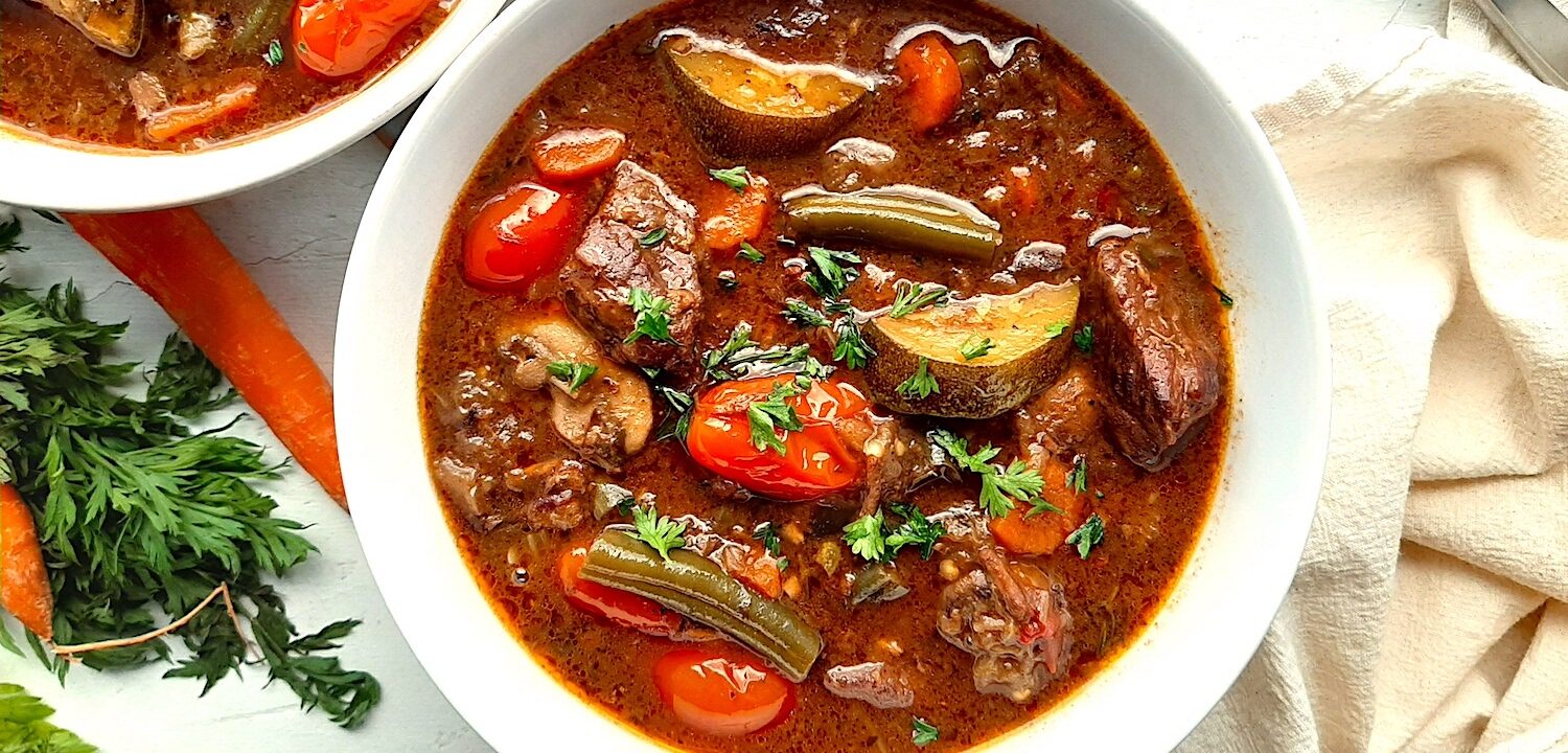 Classic stovetop beef stew with beef cubes, zucchini, carrots, tomatoes, green beans, mushrooms, and eggplant in a rich red wine broth. All served in a white bowl. A second white bowl to one side. Fresh carrots and carrot tops, a silver spoon, and a linen cloth to one side. All on a white background, overhead shot. Sugar with Spice Blog.