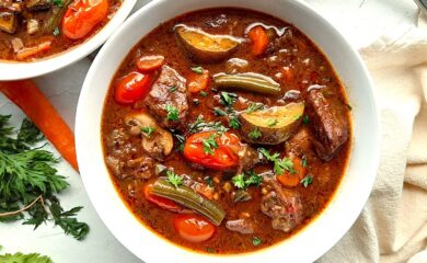 Classic stovetop beef stew with beef cubes, zucchini, carrots, tomatoes, green beans, mushrooms, and eggplant in a rich red wine broth. All served in a white bowl. A second white bowl to one side. Fresh carrots and carrot tops, a silver spoon, and a linen cloth to one side. All on a white background, overhead shot. Sugar with Spice Blog.