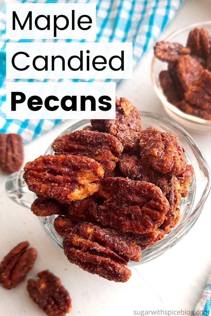 Maple candied pecans in a small glass pitcher with more maple candied pecans in a pyrex bowl behind. Blue checked dish towels nearby. A few maple candied pecans scattered on the white background. Text overlay says maple candied pecans. Pinterest Image. Sugar with Spice Blog.