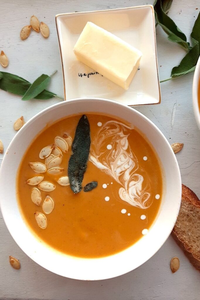 Brown Butter butternut squash soup in a white bowl garnished with roast squash seeds, fried sage, and cream swirls. A second bowl to one side. More sage leaves, roast squash seeds scatter around the white background. Pat of butter on a cream ceramic dish and two slices of sourdough bread nearby. Overhead shot. Pinterest Image. Sugar with Spice Blog.