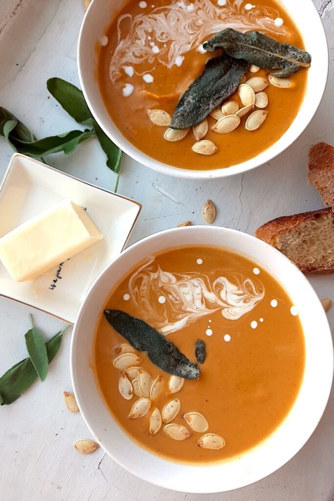 Brown Butter butternut squash soup in two white bowls garnished with roast squash seeds, fried sage, and cream swirls. More sage leaves, roast squash seeds scatter around the white background. Pat of butter on a cream ceramic dish and two slices of sourdough bread nearby. Overhead shot. Pinterest Image. Sugar with Spice Blog.